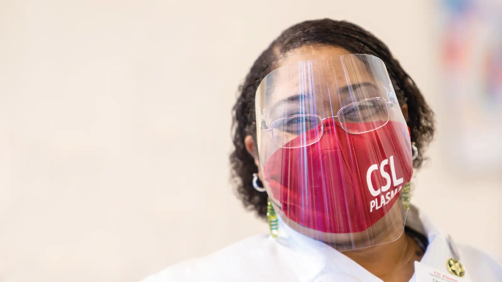 Photograph of a female CSL Plasma employee smiling while wearing a protective face shield and red face mask. This image is the cover image of CSL's 2025 Impact Report document.