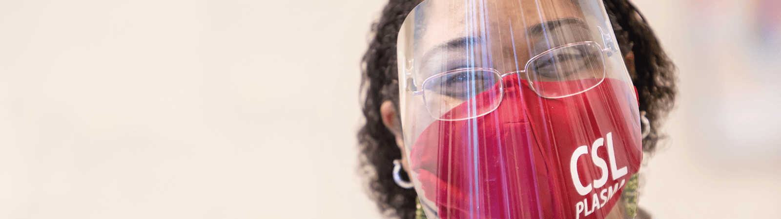 Photograph of a female CSL Plasma employee smiling while wearing a protective face shield and red face mask. This image is the cover image of CSL's 2025 Impact Report document.