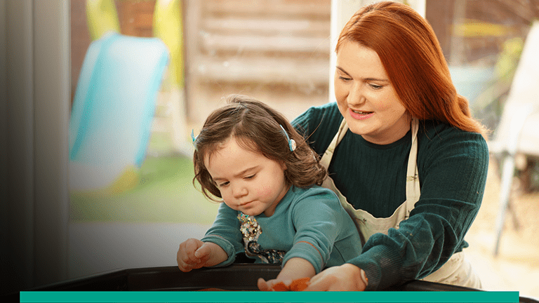 Pictured of a Red headed lady with a child on her lap. The child is playing with a toy on a table top.