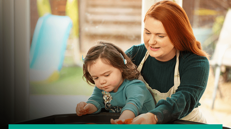 Pictured of a Red headed lady with a child on her lap. The child is playing with a toy on a table top.