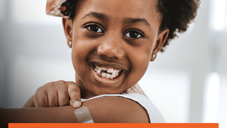 Picture of a young girl who is smiling while pointing to a band aid on her upper arm