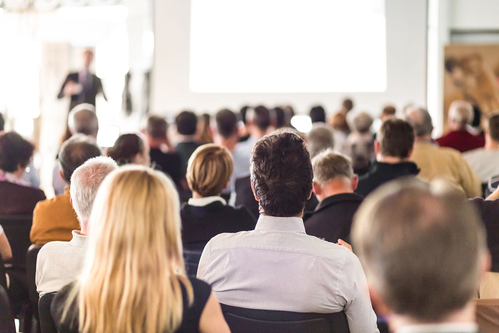 group of attendees facing the front of a room where a person is speaking  