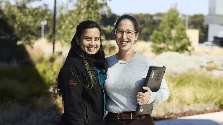Two female employees standing outdoors, smiling and have their arms around one another in a friendly embrace