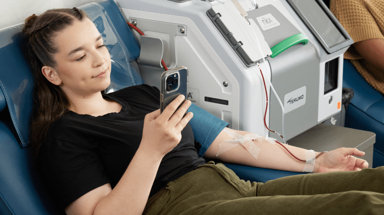 Picture of a young lady sitting in a plasma donation chair while looking at her mobile phone