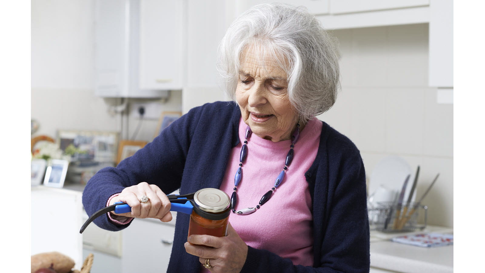 a gray haired older woman uses a tool to help open a jar