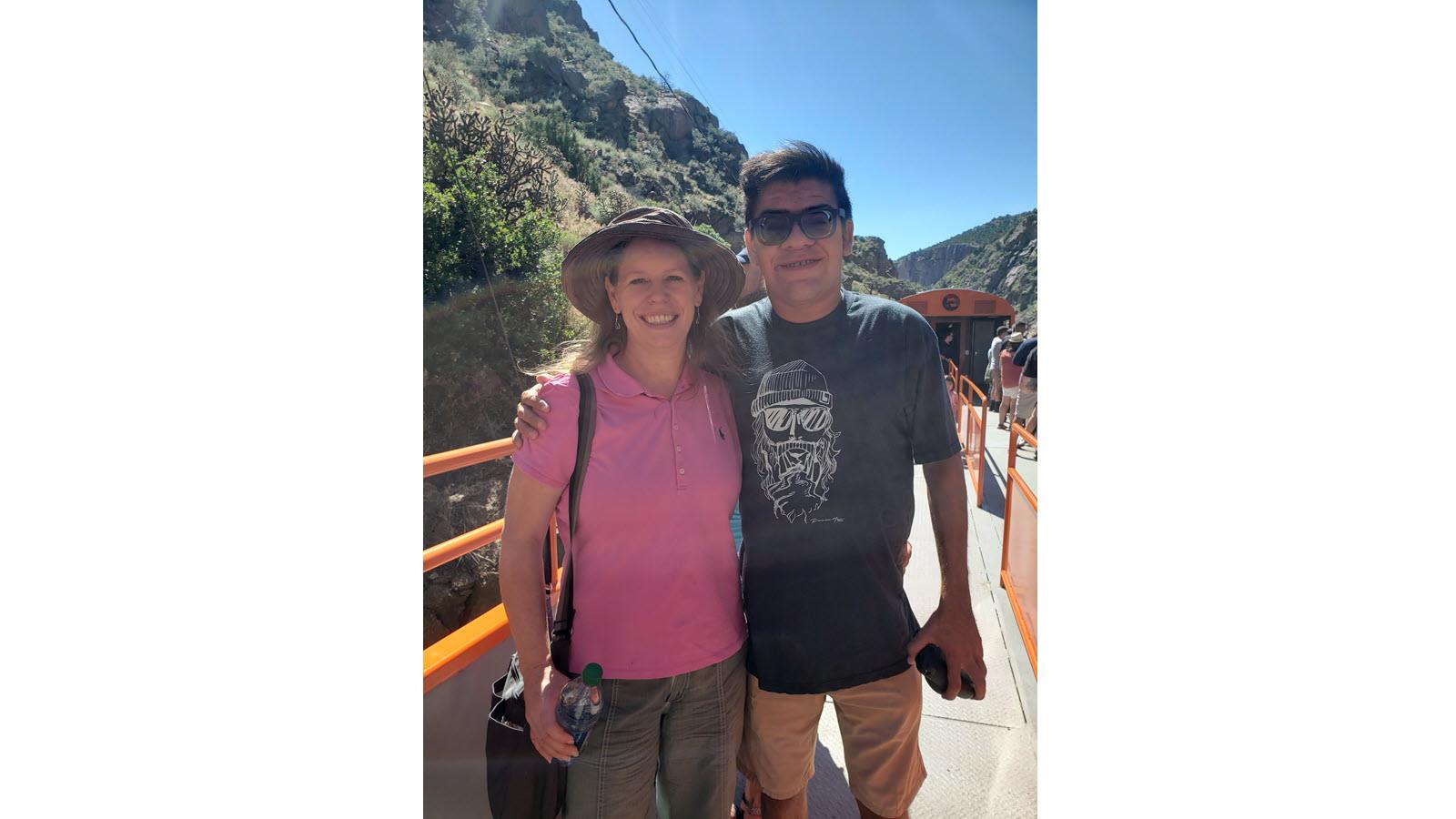 Evren in a black shirt and sunglasses with arm around mom, Kara, in a pink shit and hat outdoors in front of a mountain