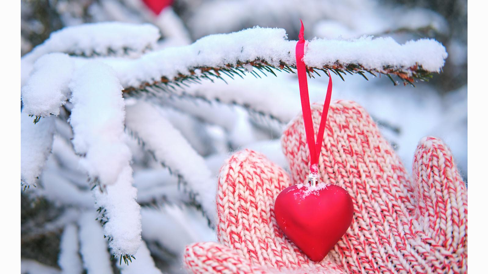 a mittened hand holds a heart hanging from a snowy Christmas tree