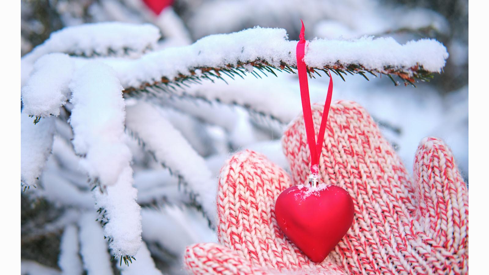 a mittened hand holds a heart hanging from a snowy Christmas tree 