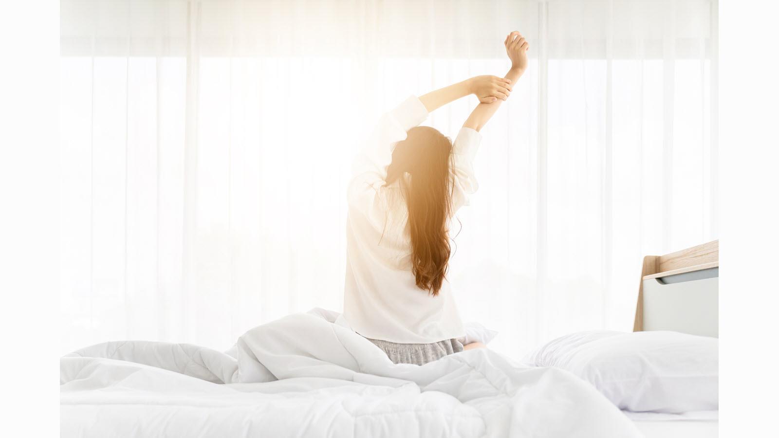 A woman takes a morning stretch in a white room with sun coming in the window.