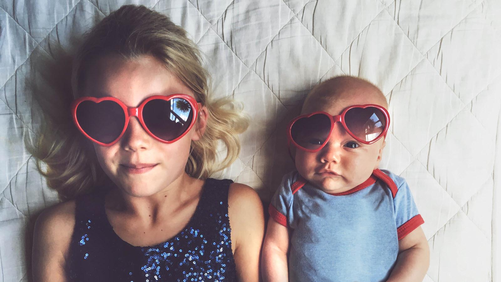baby and older sister laying on a blanket together wearing heart-shaped sunglasses