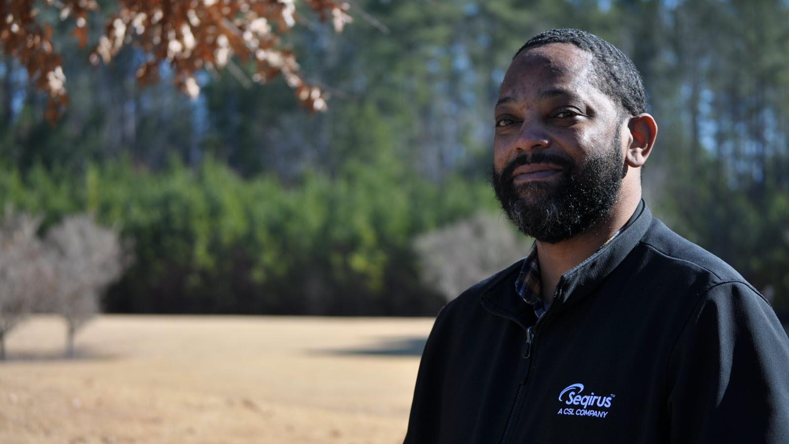 Portrait of Seqirus employee Tab Lassiter outside wearing a black polo shirt with the company logo