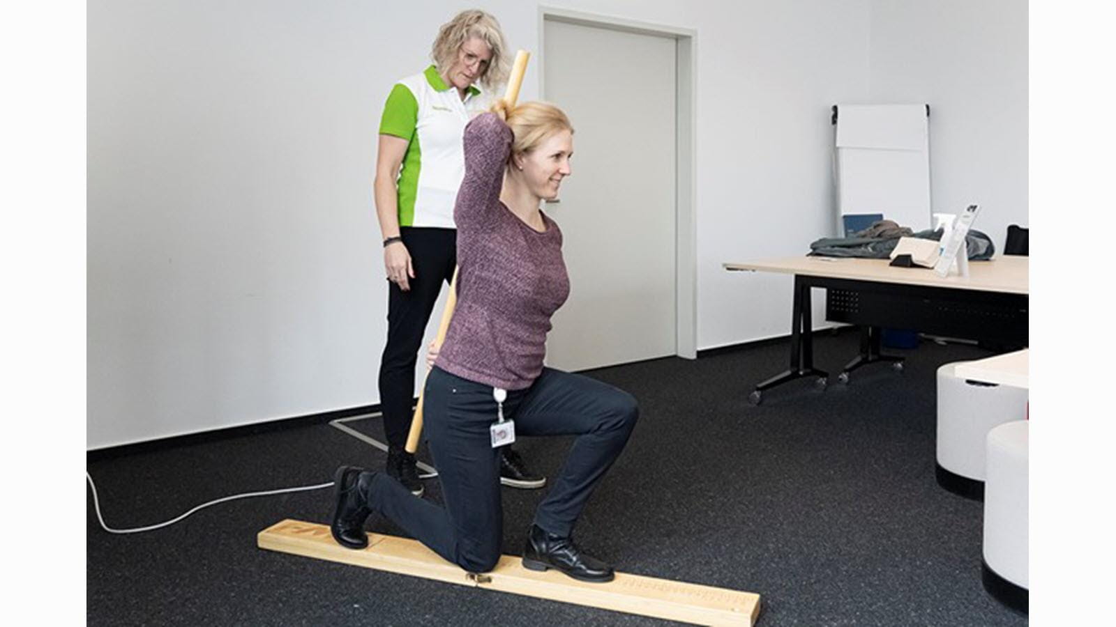 A therapist checks an employee's functional strength and flexibility while the employee is on one knee with one arm raised behind her head.