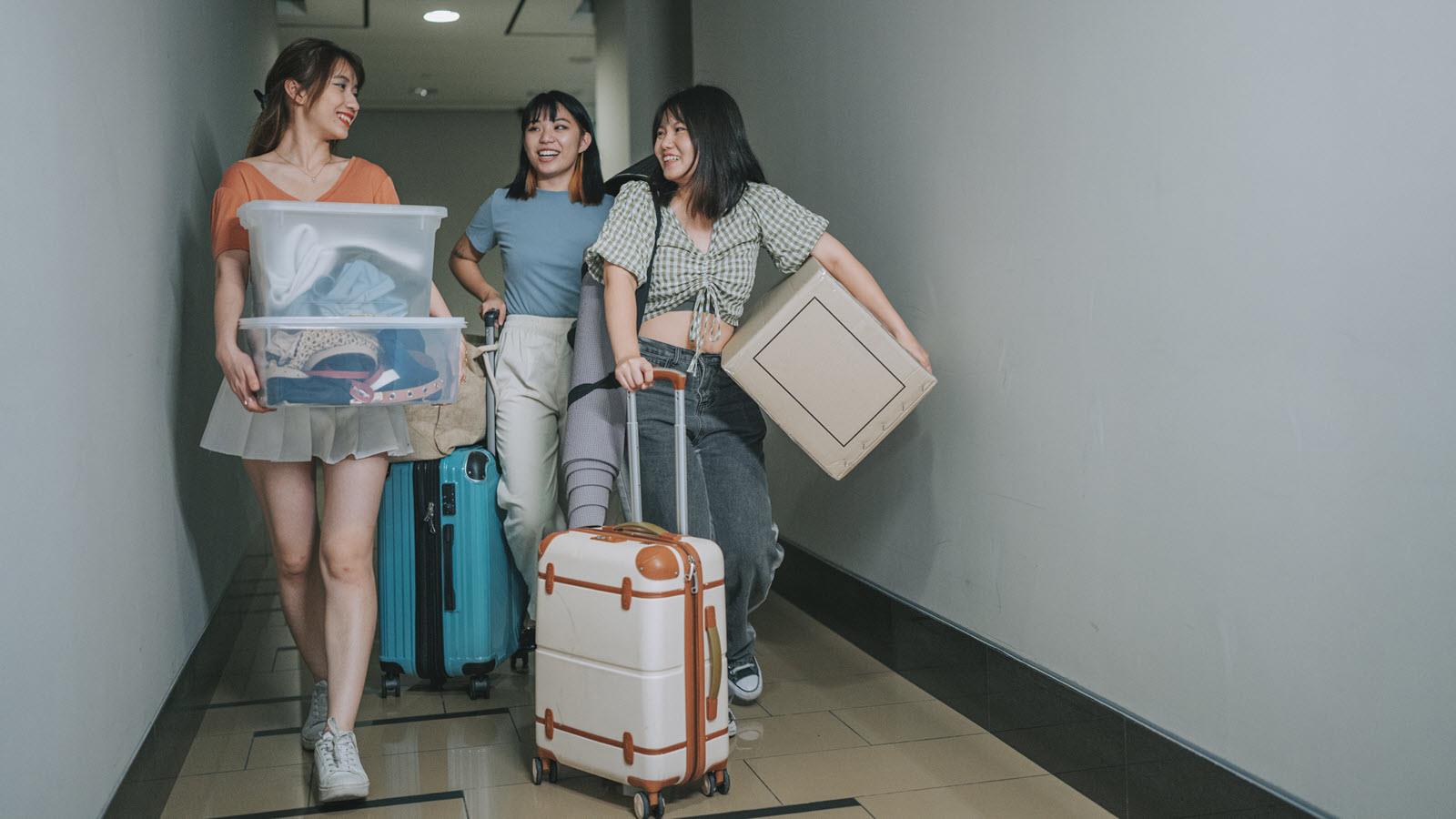 Three college students with suitcases and bins move into a dormitory.