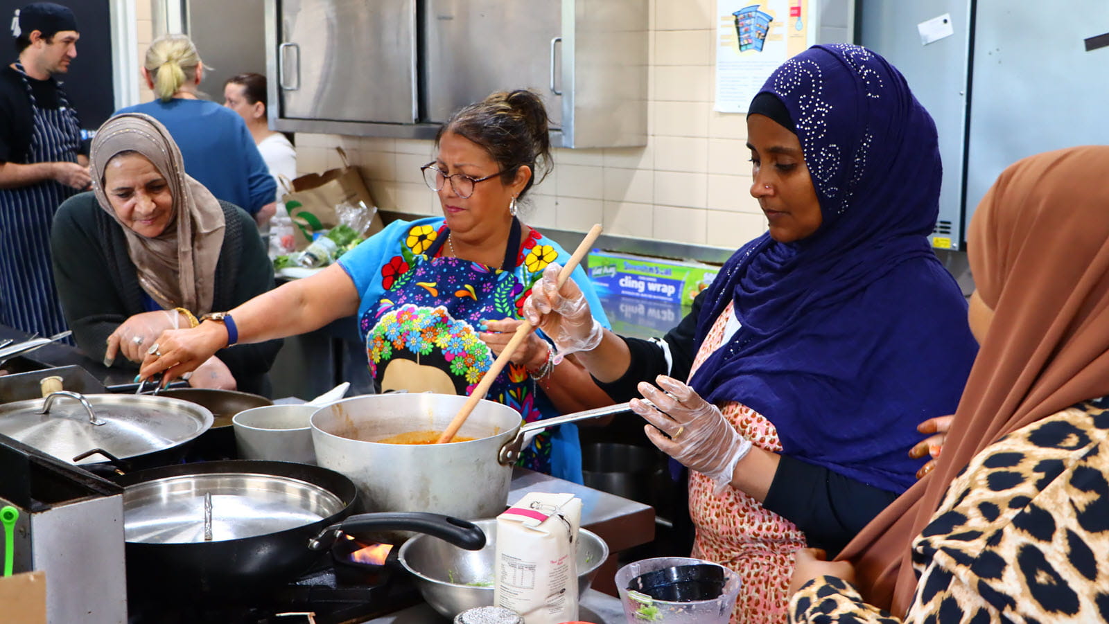 Women, including some in head scarves, prepare food on a stove together.